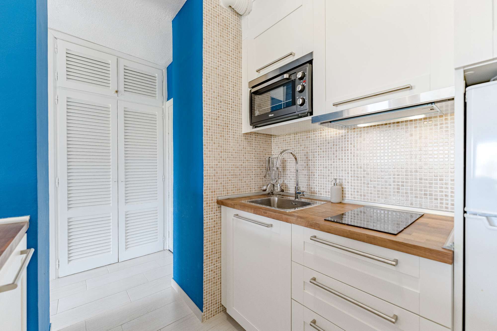 Une cuisine compacte avec des meubles blancs, un mur d’accent bleu, un four encastré et un évier en acier inoxydable.