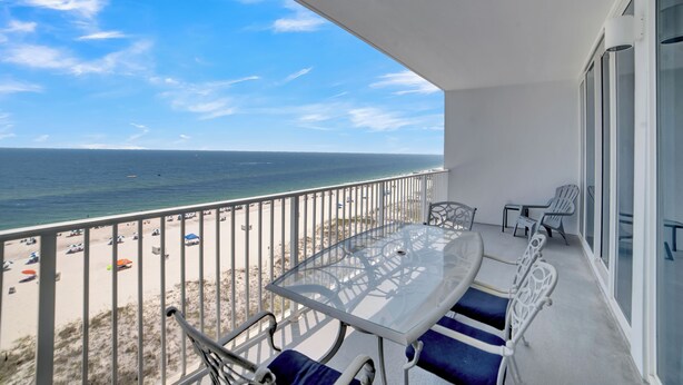 A balcony with a glass table and chairs, overlooking a beach with umbrellas and a clear sky. - Lighthouse 810 by Liquid Life (Gulf Shores)