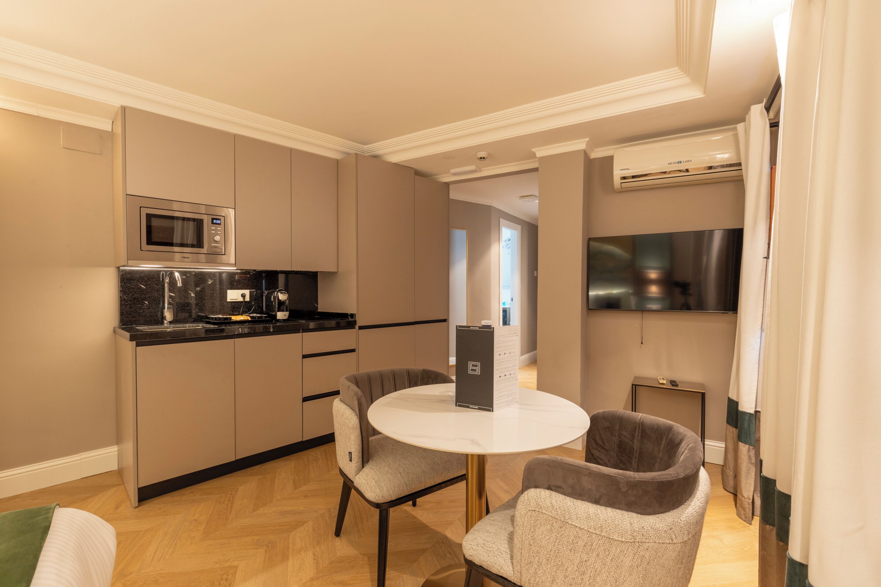 Cocina compacta con microondas, estufa y lavamanos. Hay una mesa redonda con dos sillas y un televisor pequeño en la pared.