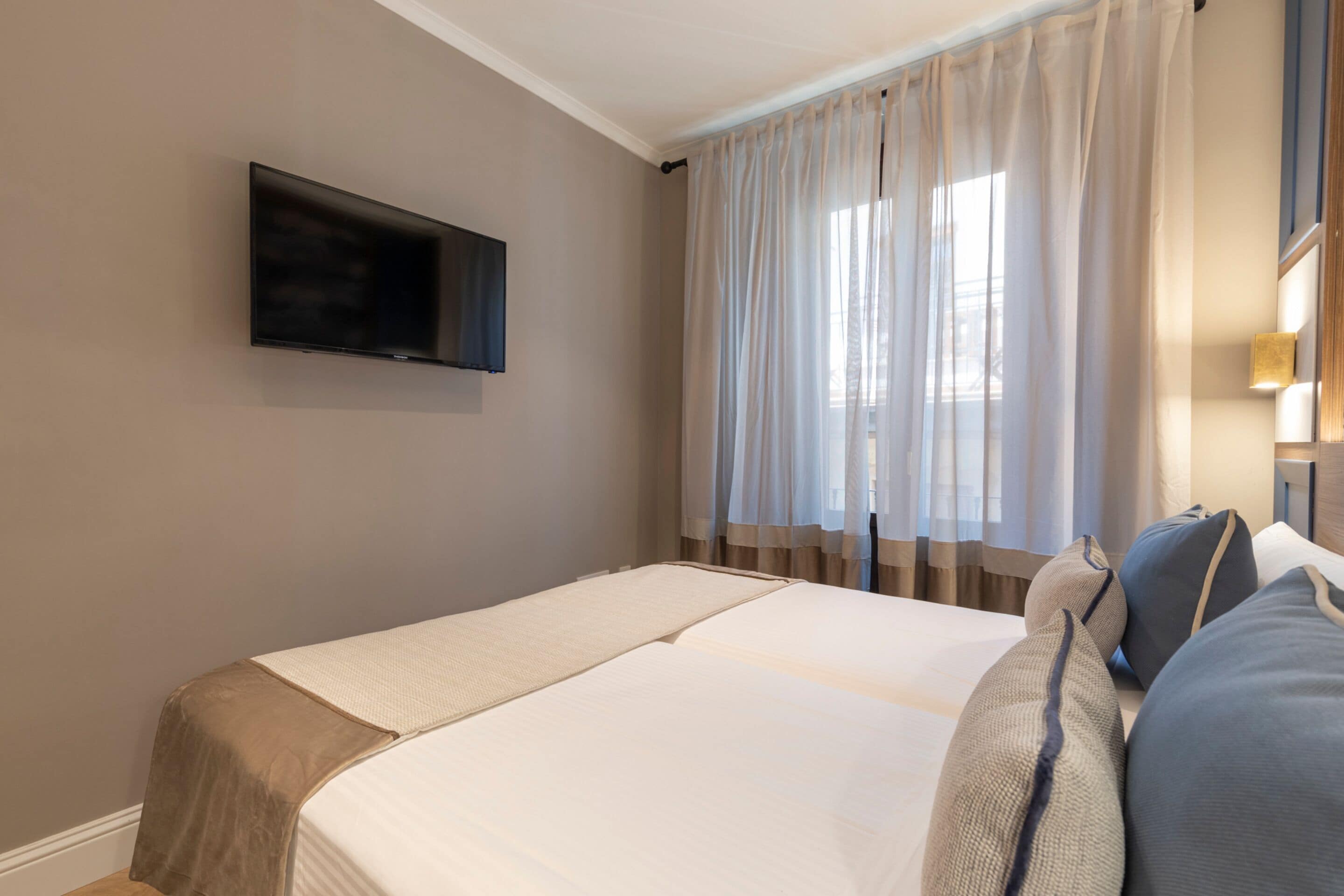 A hotel room with a bed, a flat-screen TV mounted on the wall, and a window with sheer curtains.