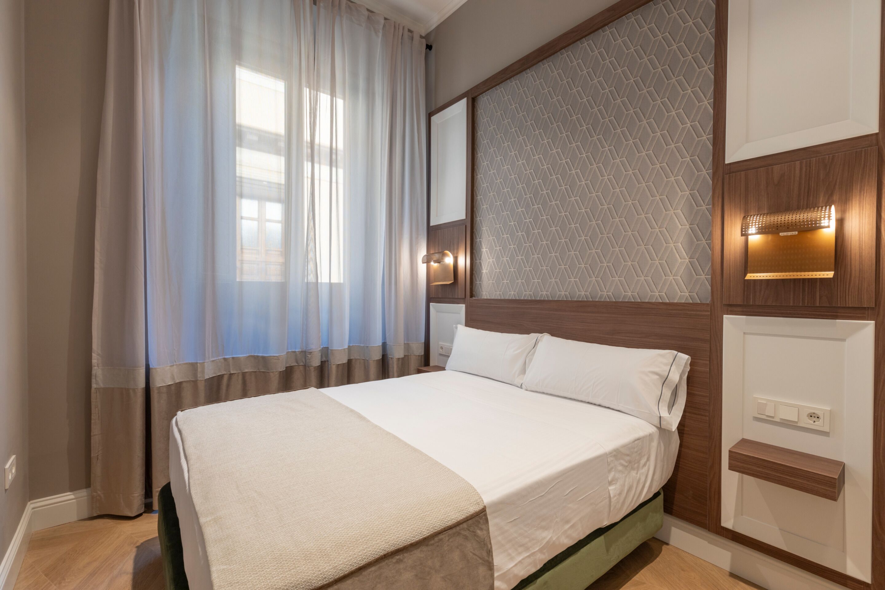 A neatly made bed with a white bedspread and a beige throw blanket, a wooden headboard with a geometric pattern, and a wall-mounted light fixture.