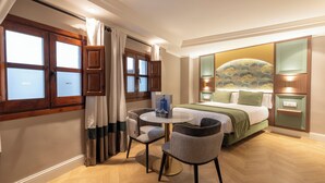Habitación de hotel con cama, una mesa redonda con dos sillas, un ventanal con cortinas y una lámpara de pared.