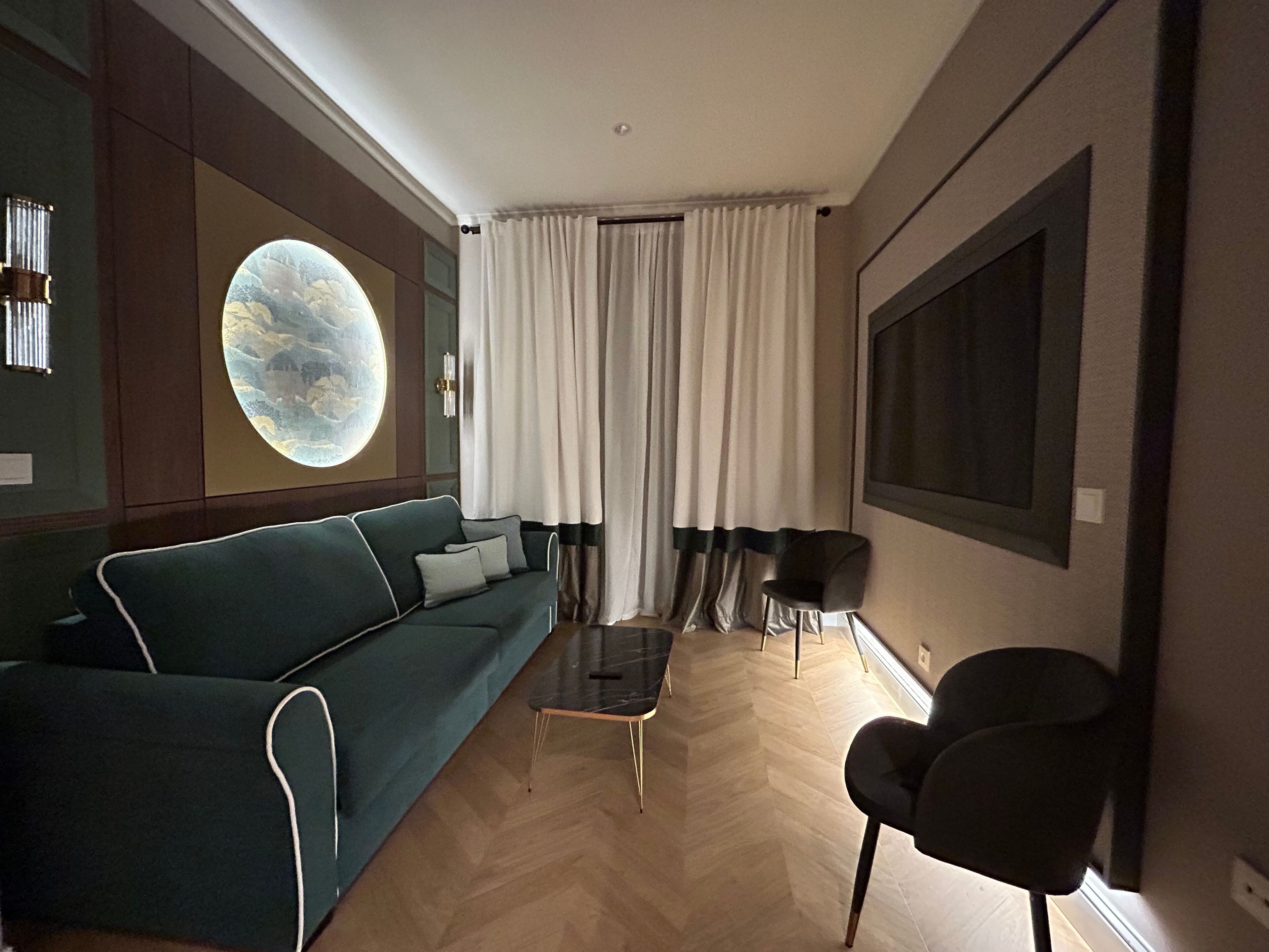 A modern living room with a green sofa, a round wall decoration, a large window with curtains, and a black chair.