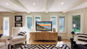 A living room with a wooden entertainment unit, a large flat-screen TV, and a view of a sunset through the windows. - Huron Shores, Hot Tub, Lakefront (Point Clark)