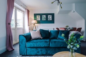 A teal sofa with cushions, a lamp, a framed plant wall, and a dining area with chairs and a table.