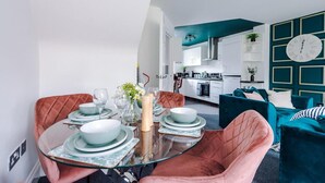 A modern living room with a glass dining table set for four, a teal accent wall, and a kitchen in the background. - Lovely 4 Bed Litherland Apartment (Liverpool)