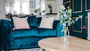 A teal velvet sofa with two pillows, a wooden coffee table with a vase, and a dark teal door with gold trim. - Lovely 4 Bed Litherland Apartment (Liverpool)