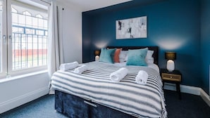 A bedroom with a bed, a nightstand, a lamp, a painting, and a window with curtains. - Lovely 4 Bed Litherland Apartment (Liverpool)