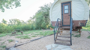 Une structure en bois couverte, dotée d’une porte et d’une échelle permettant d’y accéder, entourée d’une zone recouverte de gravier, avec une table de pique-nique et un banc.