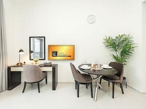 A modern hotel room with a dining area, a desk with a lamp, a large mirror, a framed sign, and a potted plant.
