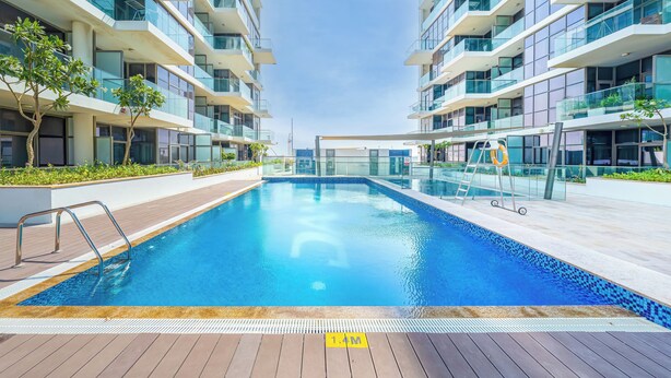 A modern swimming pool with a wooden deck, surrounded by glass balconies and a clear sky. - Luxury StayCation - Studio in Damac Hills with Balcony and Golf Views (Dubai)