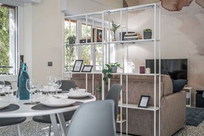 Un espace de repas moderne avec une table blanche dressée pour un repas, un canapé et une bibliothèque remplie de livres et de plantes.