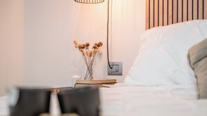 Siististi pedattu sänky, jossa on valkoinen peitto ja tyyny, yöpöytä, jossa on kirja ja kynttilä, sekä seinään kiinnitetty valaisin.