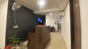 Un espace d’accueil avec un mur sombre, un comptoir de réception en bois, un téléviseur fixé au mur, un extincteur et une plante en pot.