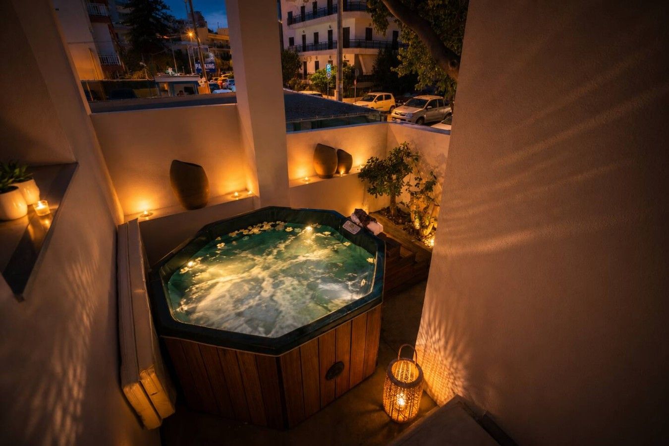 A hot tub with a wooden exterior, surrounded by candles and a lantern, set against a building facade.