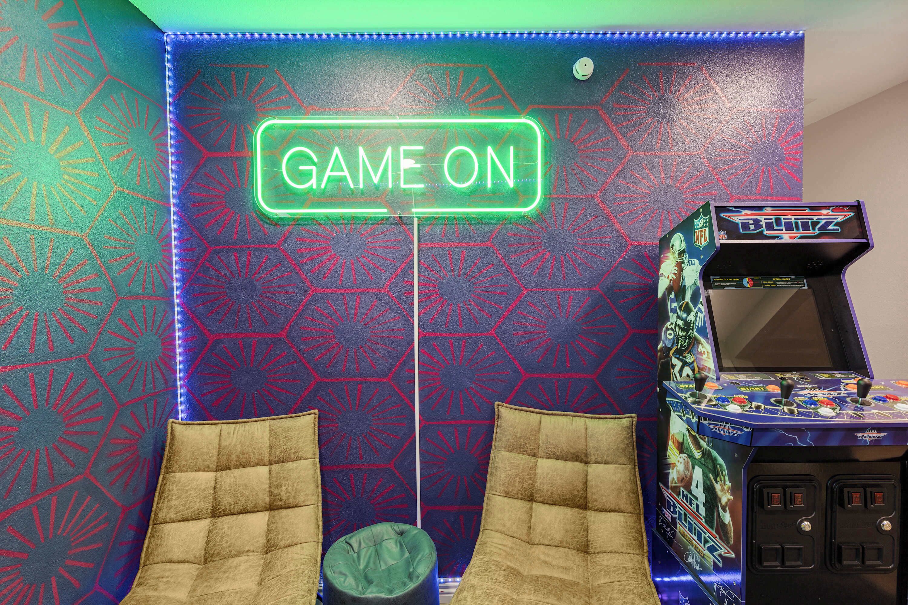A neon 'GAME ON' sign, two cushioned chairs, and a video game arcade machine.