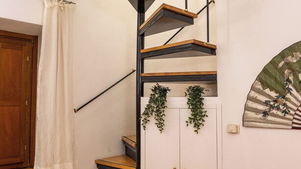 Una escalera con escalones de madera y barandales negros, un sofá rojo con un cojín azul y una planta colgante en la pared.