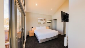 A bedroom with a large bed, a bedside table, a TV mounted on the wall, and a window with a view of trees. - The Staggerinn - Dinner Plain (Dinner Plain)