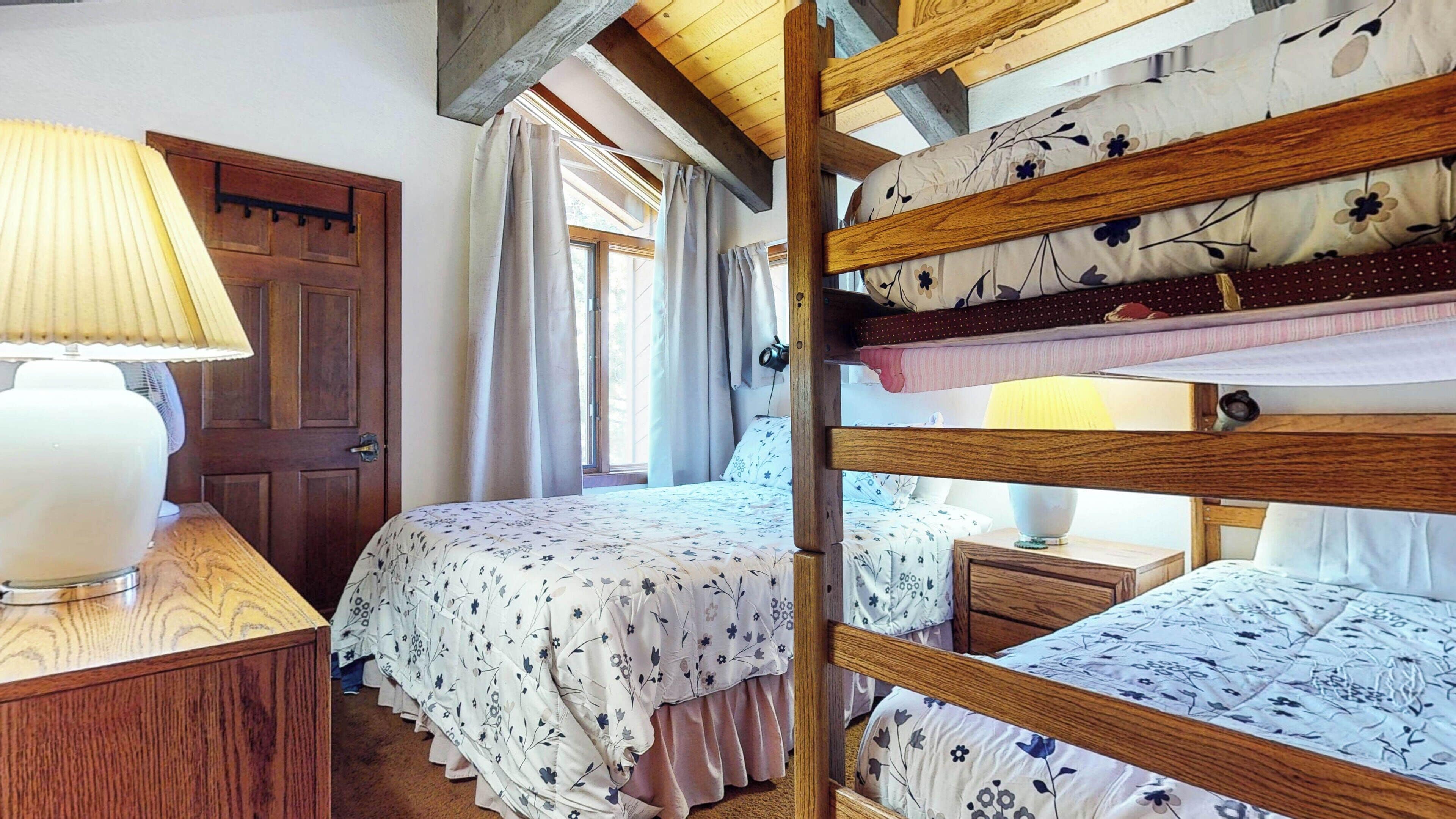 Un dormitorio con una cama litera de madera, una mesita de noche con lámpara, una ventana con cortinas y una puerta.