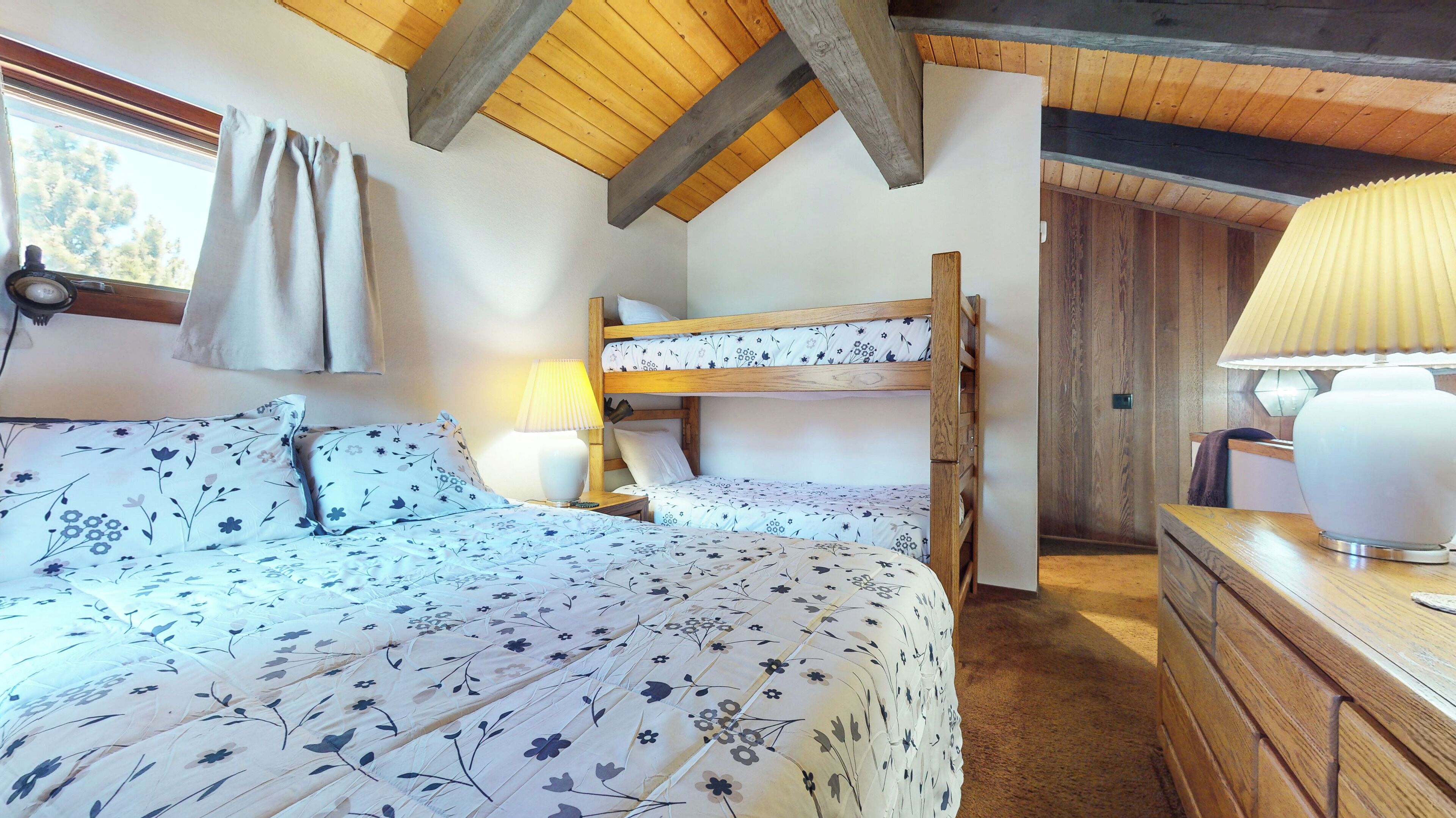 A bunk bed with floral bedding, a bedside lamp, and a wooden dresser in a room with exposed wooden beams and a window with curtains.