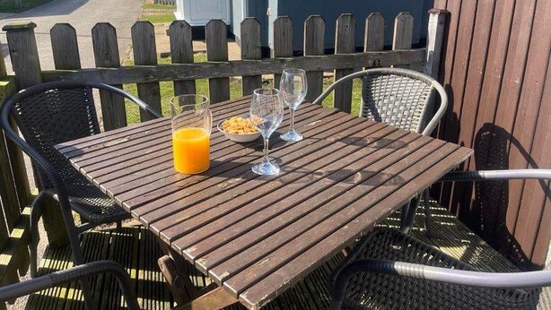A wooden table with two chairs, a glass of orange juice, and a bowl of cereal on a balcony with a wooden fence and houses in the background. - Seaside Escape Chalet in Bridlington South Shore (Bridlington)