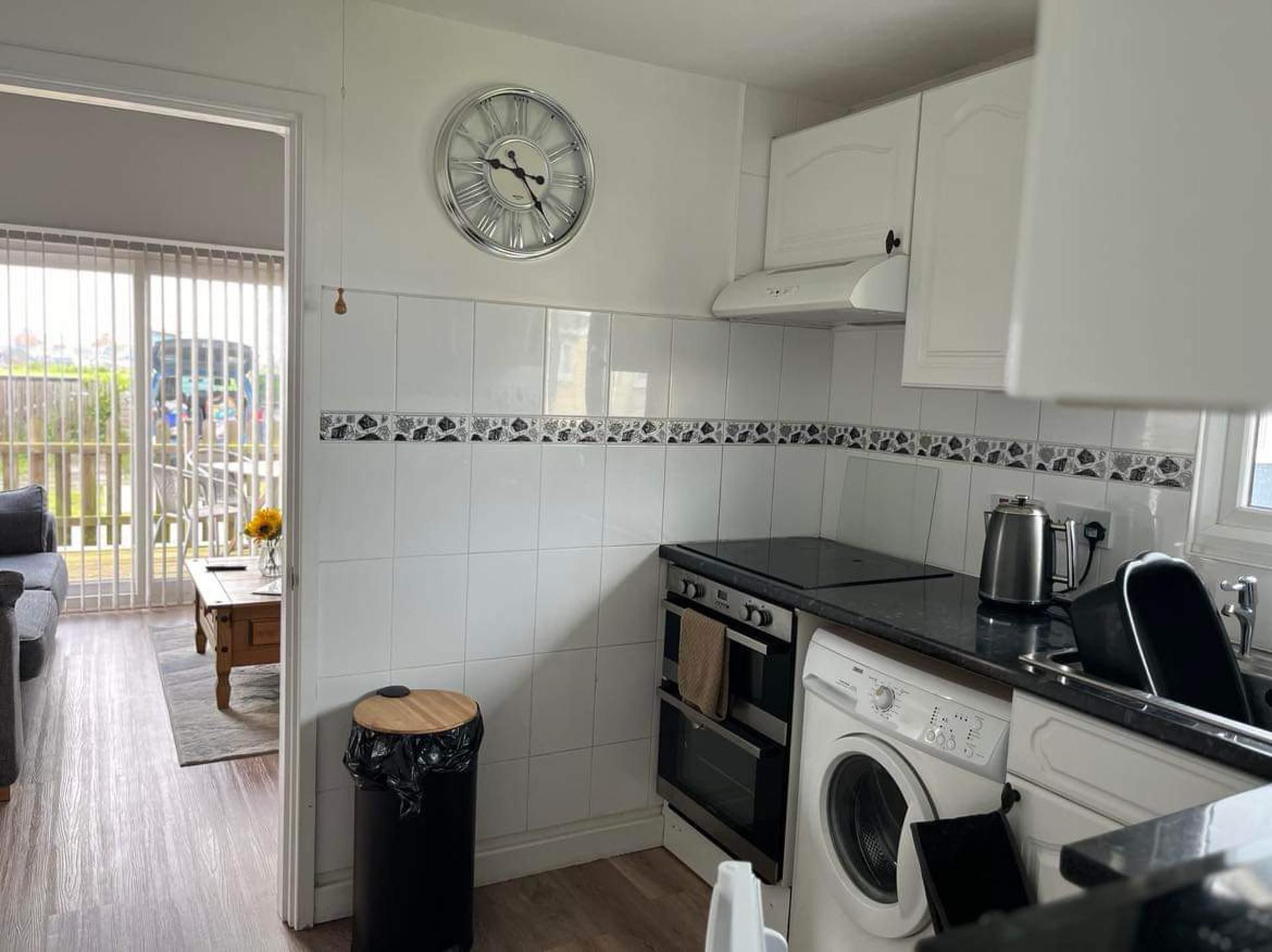 A compact kitchen with white cabinets, a black countertop, a built-in oven, a washing machine, and a clock on the wall.