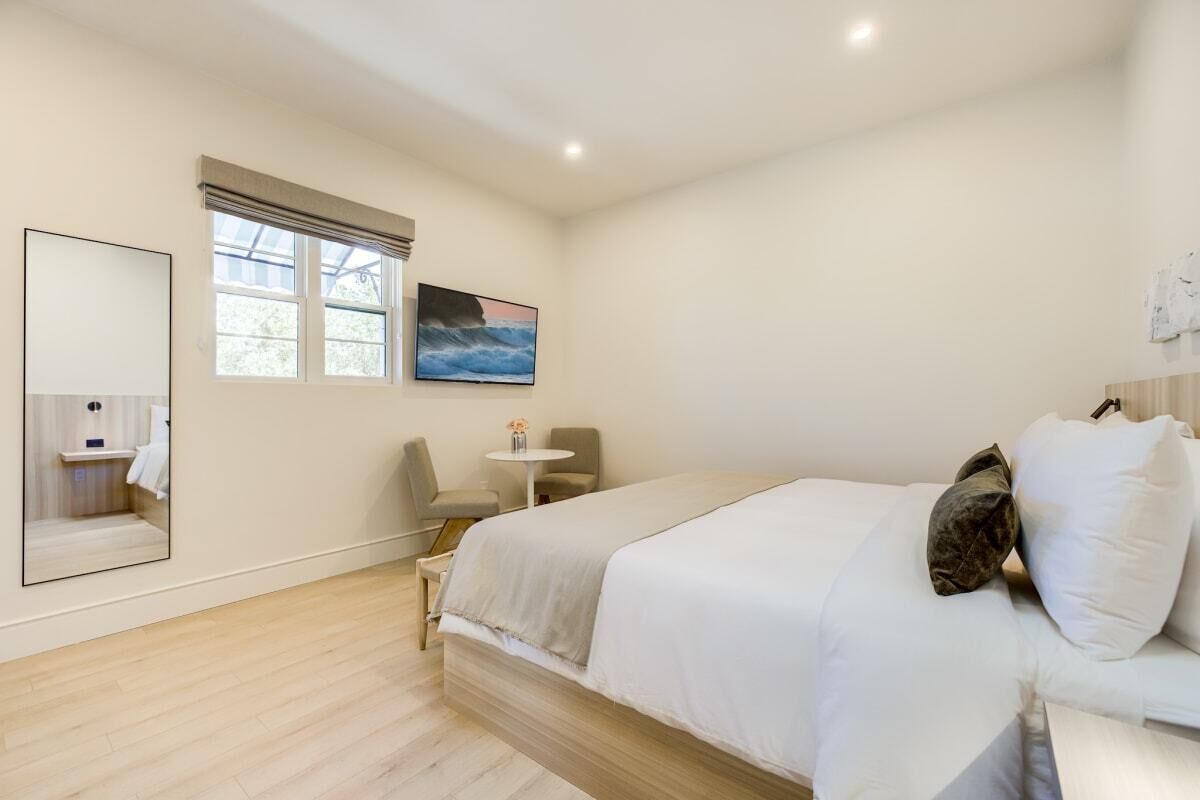 A bedroom with a bed, a small table, a chair, a window with curtains, a painting, and a mirror.