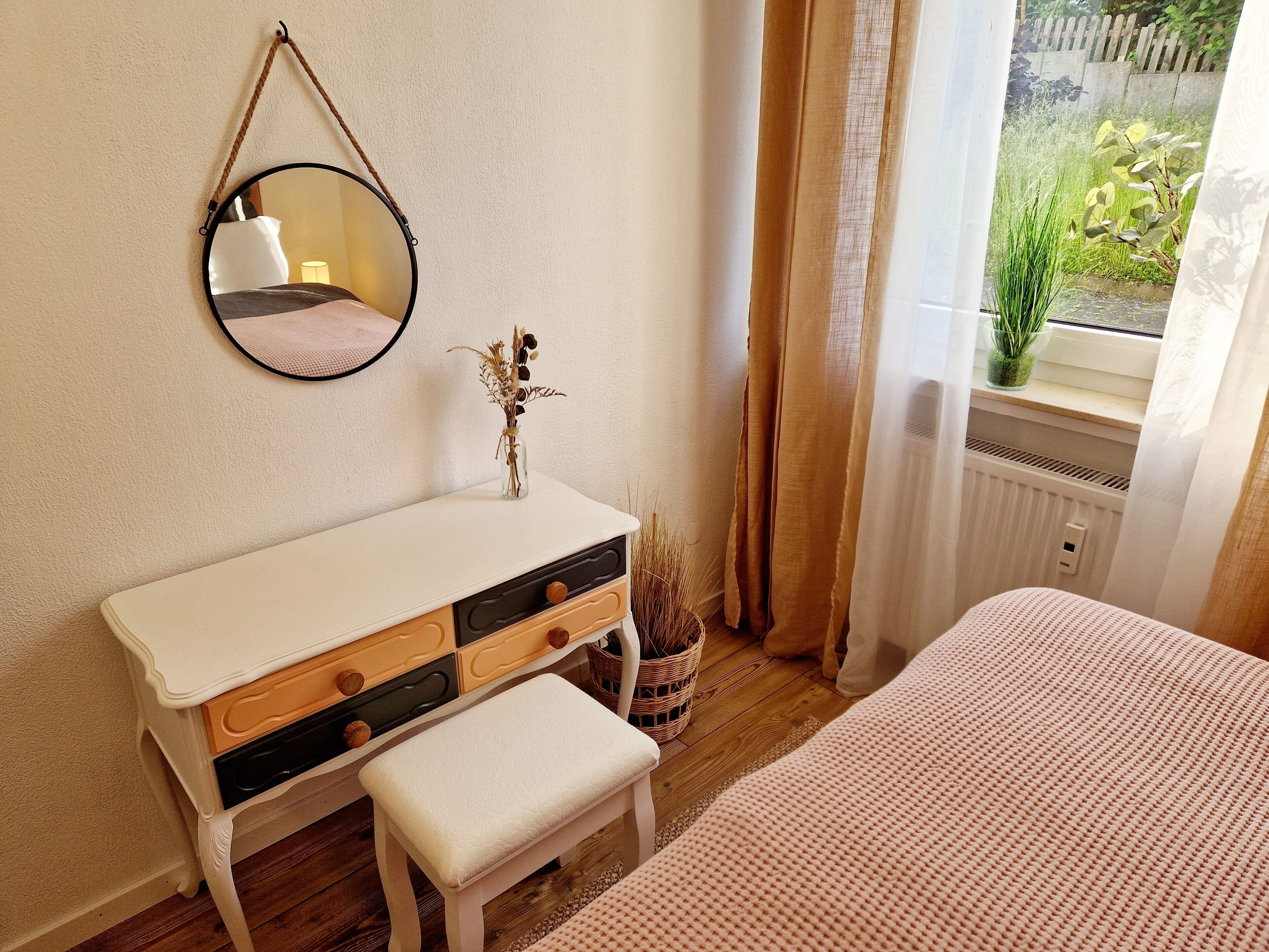 A bedroom with a white dresser, a mirror, a bench, a window with curtains, and a view of greenery outside.