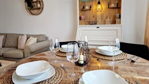 Un coin repas avec une table en bois dressée pour quatre personnes, entourée de sets de table en osier, d’assiettes blanches et de verres à vin. On peut également apercevoir un mur décoratif avec une enseigne indiquant « café » et des étagères remplies d’ustensiles de cuisine.