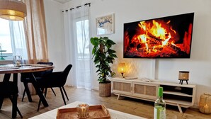 Un salon moderne avec un téléviseur diffusant une image de feu de cheminée, un coin repas avec des chaises, une table avec des verres et une bougie, et une plante.