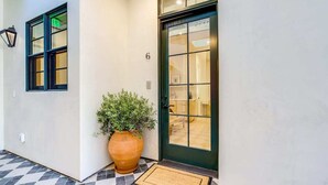 Un pasillo con piso a cuadros, una planta grande en maceta, una puerta con panel de vidrio y el número 6 en la pared.