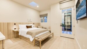 A modern bedroom with a large bed, a bench, a nightstand, and a view of the outside through a glass door.