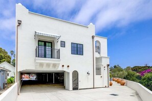 Un edificio blanco de dos pisos con cochera cubierta, un balcón y una estructura pequeña adosada.