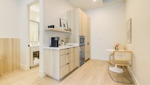Una cocina moderna con armarios de madera clara, un refrigerador de acero inoxidable y una cafetera. Hay un pequeño comedor con una silla vintage y una mesa blanca.
