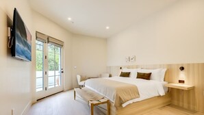 Un dormitorio con una cama grande, un banco, un televisor montado en la pared y una ventana con persianas.