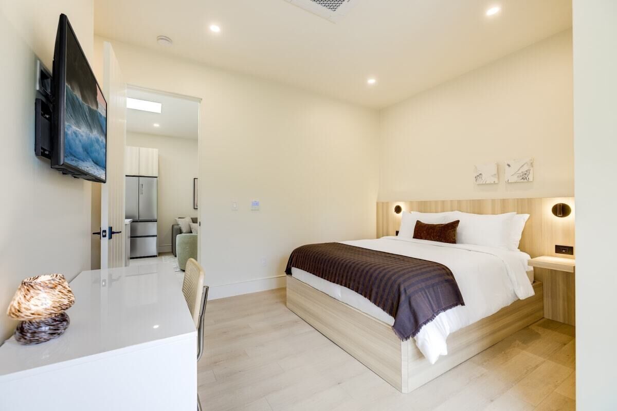 Quarto de hotel moderno com cama, mesas de cabeceira, televisão de tela plana, escrivaninha com abajur e vista para o corredor.