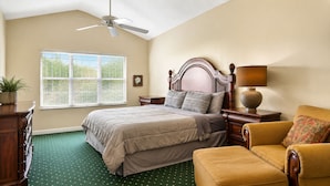 Quarto com cama, cadeira, mesa de cabeceira, luminária, ventoinha no teto e janela com persianas.