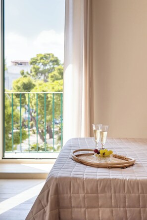 A bed with a beige quilted bedspread, a tray with glasses and fruit, and a view of a balcony with greenery and a building in the background. - Viesthouse (Vieste)