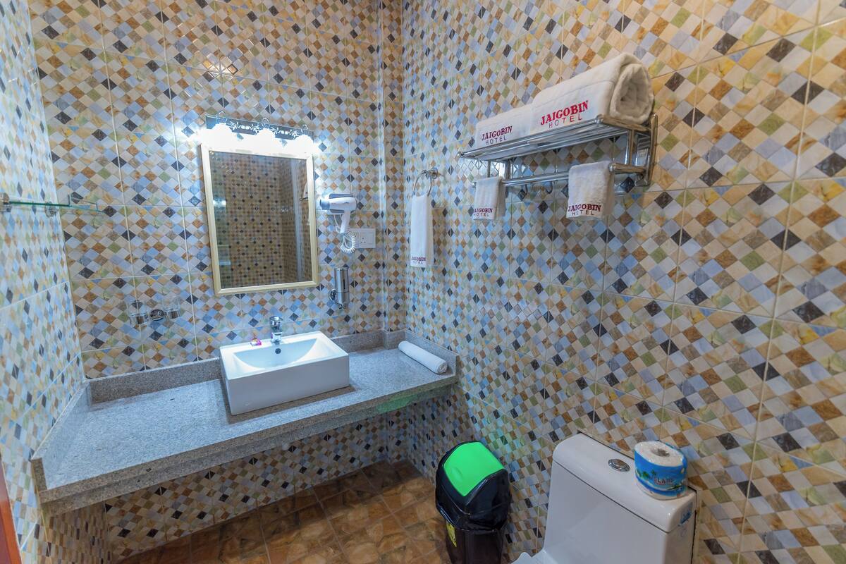 A bathroom with a patterned tile wall, a sink, a mirror, a towel rack with towels, and a trash bin.
