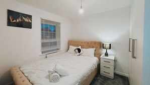 A neatly made bed with a white duvet, a bedside lamp, and a small bedside table with a lamp and a candle. - Cozy Windmill House in Smethwick (Smethwick)