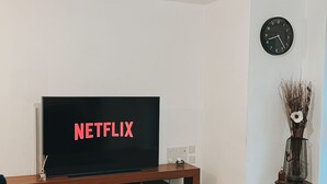 A living room with a TV displaying 'NETFLIX' in red letters, a clock on the wall, and a decorative vase with branches. - Cozy Windmill House in Smethwick (Smethwick)