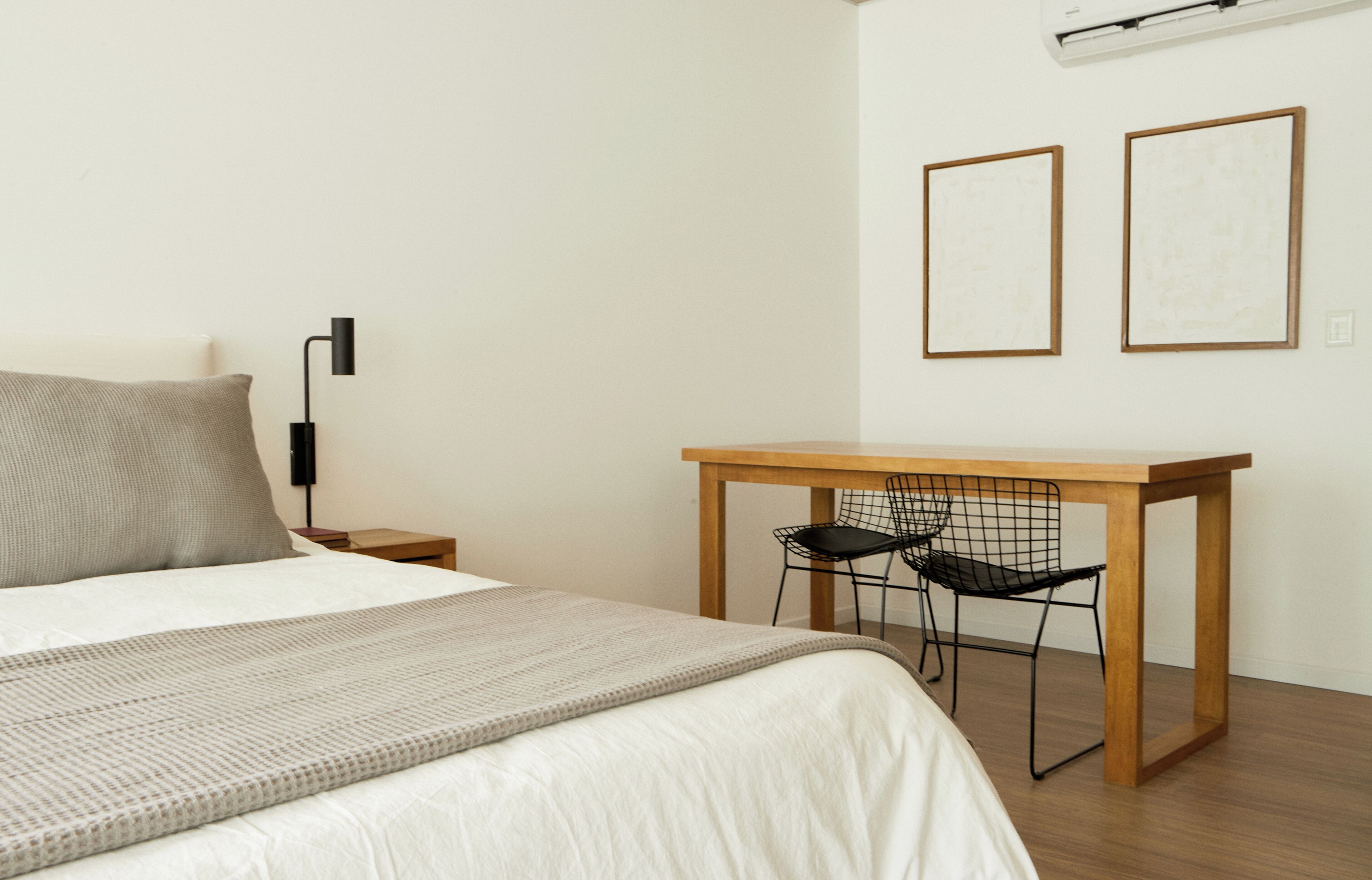 Un dormitorio con cama, mesita de noche, lámpara, escritorio, dos sillas y dos cuadros en la pared.