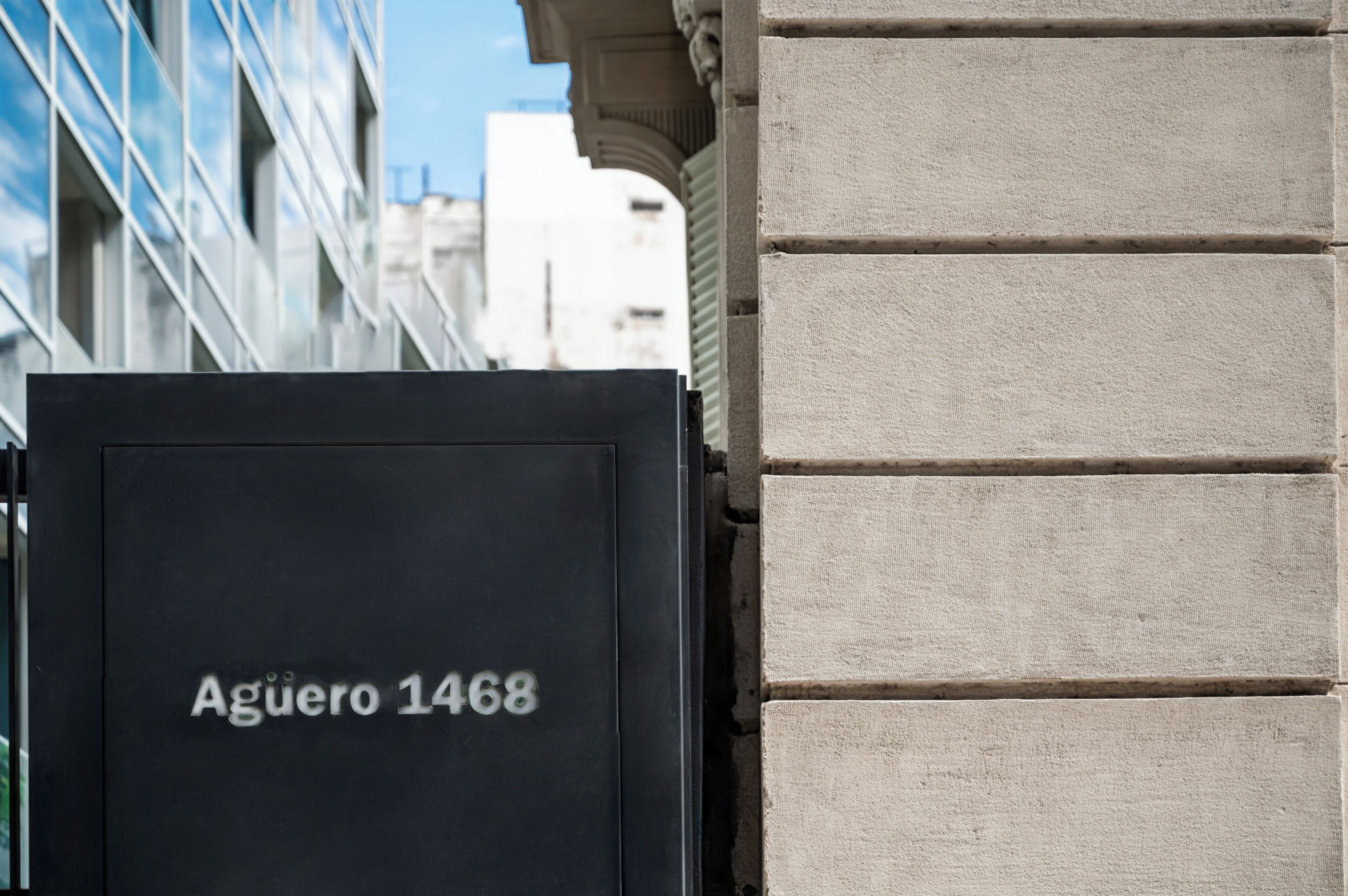 A black mailbox with "Aguero 1468" written on it, attached to a building with a stone facade and a modern glass structure in the background.