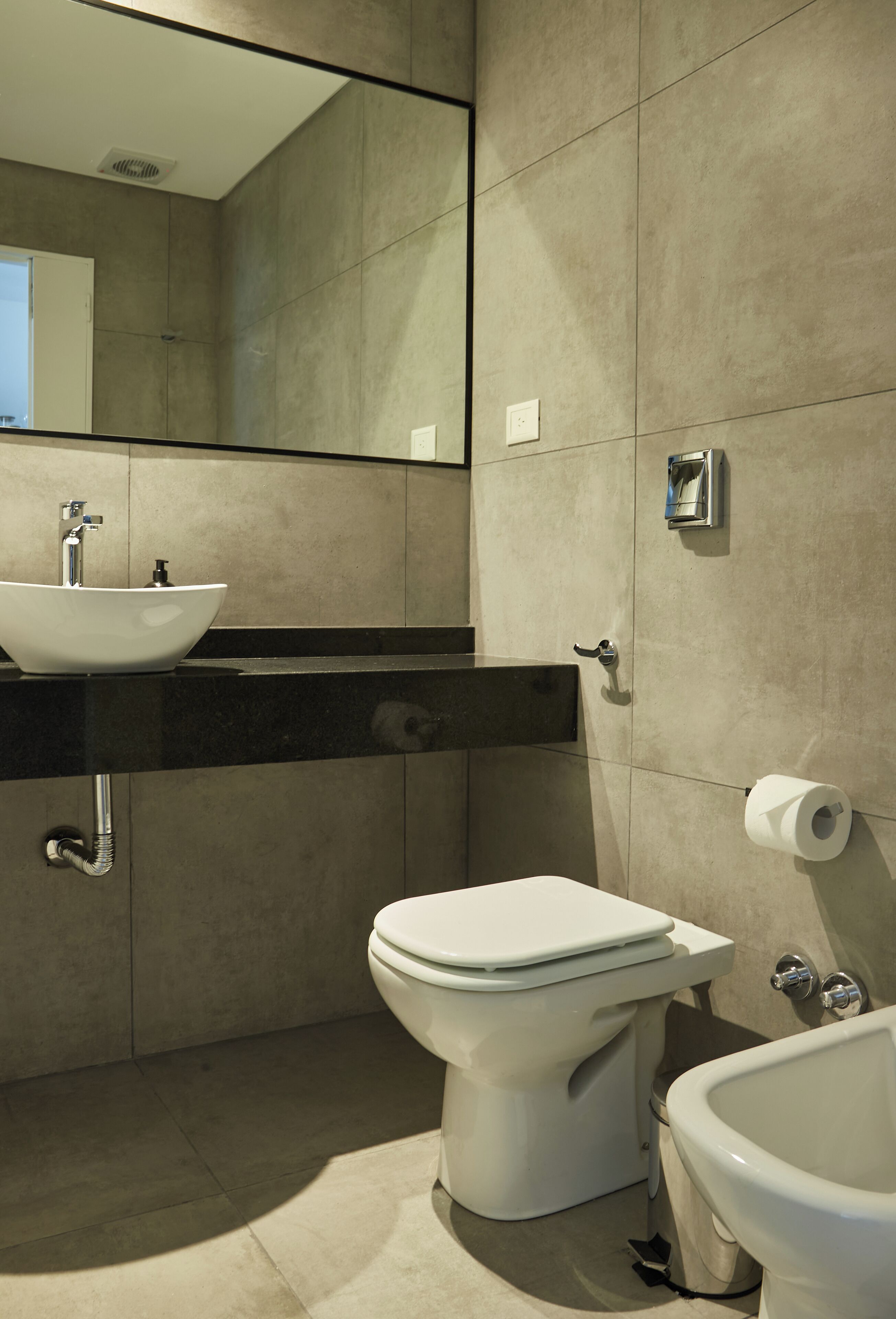 Un baño moderno con lavamanos, inodoro y bidé blancos, un espejo grande y un dispensador de toallas.