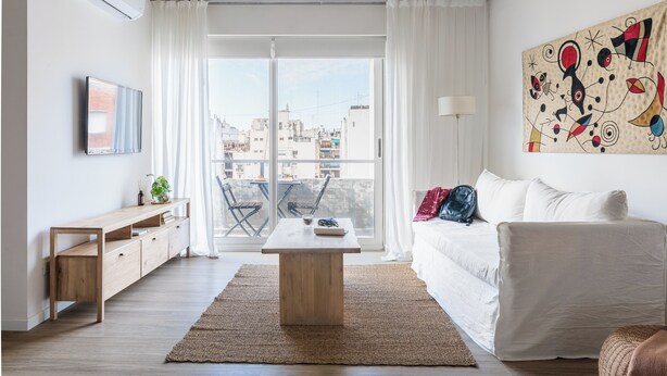 Un salon moderne avec une table basse en bois, un canapé blanc, un téléviseur fixé au mur et une grande fenêtre donnant sur des immeubles.