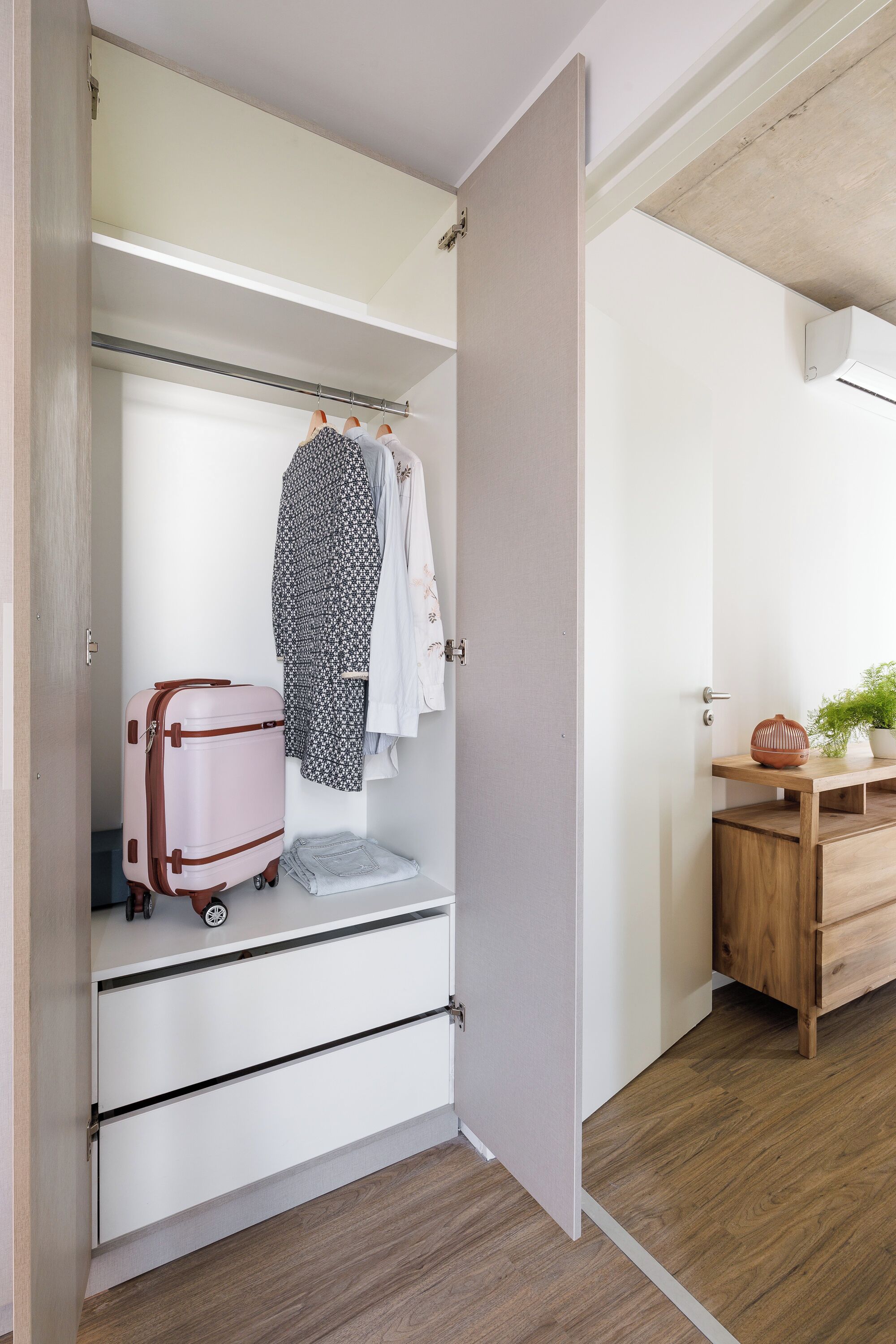 Une pièce avec un plancher en bois, une armoire blanche avec une tringle à vêtements, une valise et une petite table en bois sur laquelle est posée une plante.