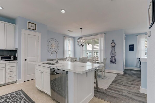 A modern kitchen with white cabinetry, a central island, and a dining area with a chandelier. - Beachy Keen-New Ocean Side Cottage- Now Pet Friendly! 3BD/3.5 BA. Sleeps 8 (Galveston)