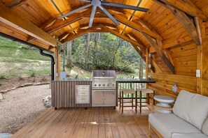 Un espace cuisine extérieur comprenant un barbecue, une terrasse en bois et un coin salon avec un canapé et des fauteuils.