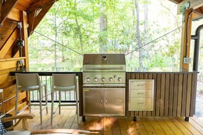 An outdoor kitchen with a gas stove, four burners, and a stainless steel cabinet with drawers. There are two chairs and a table nearby.
