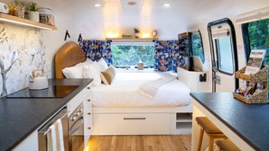 A compact, well-lit room with a bed, a small kitchenette, and a window offering a view of greenery. - Creekside Fern in Pisgah Forest (Pisgah Forest)